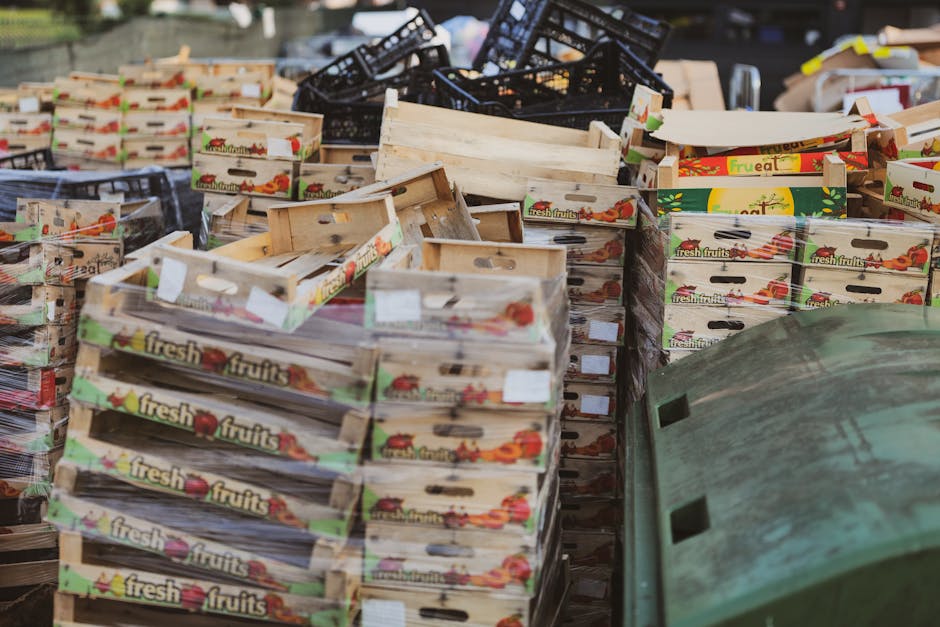 A large outdoor collection of cardboard fruit boxes stacked in various orientations, some leaning and overlapping, with a mix of new and used wooden crates featuring colorful printed labels that read 'fresh fruits.' The boxes display different finishes and textures—smooth, glossy printed areas contrasting with rough, fibrous edges of the cardboard and wood. Behind the stacks, dark plastic crates are piled haphazardly, some tilted or partially visible, while a green plastic bin with a curved rim is partially in the foreground. The surrounding environment suggests an outdoor storage or waste collection area, with natural lighting highlighting the textures and imbalance of the stacked packaging materials. This scene relates to the context of waste removal and alternative on-site clearance services for commercial or retail outlets, emphasizing the volume and packaging materials typical in rubbish collection for retail waste management. Waste Removal Colliers Wood occasionally handles such bulk waste in accordance with private disposal standards, supporting effective rubbish clearance for businesses.