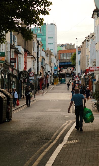 A busy urban street scene on a commercial shopping street, with pedestrians walking along the pavement on both sides and across the crosswalk. The foreground features a man wearing a blue shirt holding a large green plastic bag filled with waste, walking away from the camera towards the street. On the left, there are small shopfronts with various signs, some with awnings, and a few people standing outside or entering shops. The right side shows more shops and a few pedestrians, with some displayed merchandise or signage visible. In the background, higher-rise buildings with glass and concrete facades dominate the skyline, and a blue delivery van is parked further along the street. The environmental atmosphere appears daytime with natural lighting and an overcast sky, with indications of ongoing activity. This scene illustrates typical street-level activity in a commercial area, aligning with the context of independent waste collection or private rubbish removal services conducted on-site or in public spaces, as seen through the man carrying waste for disposal or collection by companies like Waste Removal Colliers Wood.