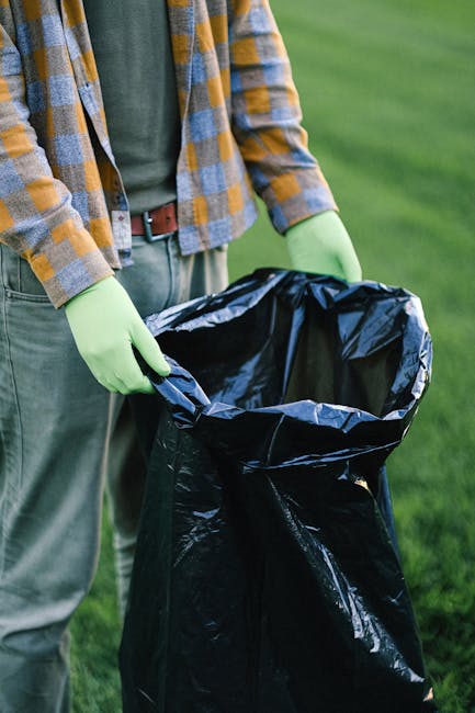 A person wearing a plaid shirt with shades of orange, grey, and yellow, paired with grey trousers and a brown belt, is holding open a black plastic rubbish bag lined with a shiny silver inner layer. The person is also wearing light green gloves, and their hands are positioned around the top edge of the bag, which is partially filled with waste. The setting appears to be outdoors on a grassy area with vibrant green grass visible in the background, suggesting a clean-up activity or ongoing waste disposal. The image highlights a moment of on-site rubbish collection, which aligns with private waste handling services provided by Waste Removal Colliers Wood, emphasizing the step of manual rubbish collection that may complement or offer an alternative to traditional council refuse services.
