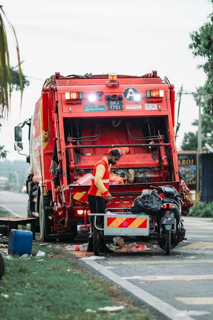 A waste collection worker wearing a high-visibility yellow and red uniform stands at the rear of a large, red garbage truck with an open waste compactor. The truck is parked on the side of a street, with its rear loading mechanism lifted and partially filled with waste. The worker appears to be managing or inspecting black plastic trash bags placed on a small metal platform attached to the truck. The environment features a paved roadway with a curb and some greenery on the side, including a small section of grass and trees in the background. Nearby is a motorbike parked close to the truck. The scene is illuminated by natural daylight, with a slightly overcast sky. This setup reflects an example of on-site waste collection, commonly used for private or independent rubbish removal services, such as those offered by Waste Removal Colliers Wood, highlighting the practical aspects of waste disposal on urban streets without reliance on local authority collections.