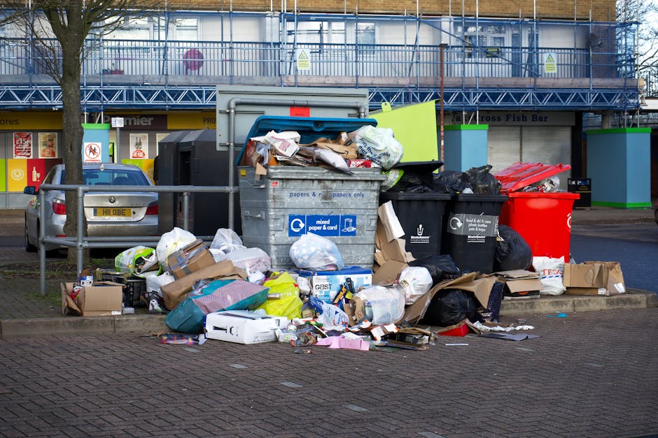 An outdoor scene showing several overflowing rubbish bins and scattered waste on a paved area adjacent to a parking lot. The central grey bin is open at the top, filled with mixed paper, cardboard, and plastic waste. To its right, there are black and red bins, with black bin bags and loose trash spilling onto the ground, including cardboard boxes, plastic packaging, and paper waste. The scene is set in front of a commercial building with a blue scaffolding structure on the upper level and storefronts with yellow signage below, indicating retail shops or services. On the left, a parked silver car is visible behind a metal railing, with a leafless tree nearby. The environment suggests a public space where waste has accumulated, highlighting the importance of proper rubbish collection and disposal services such as those offered by Waste Removal Colliers Wood, especially for efficient on-site clearance and alternative waste handling in urban areas.