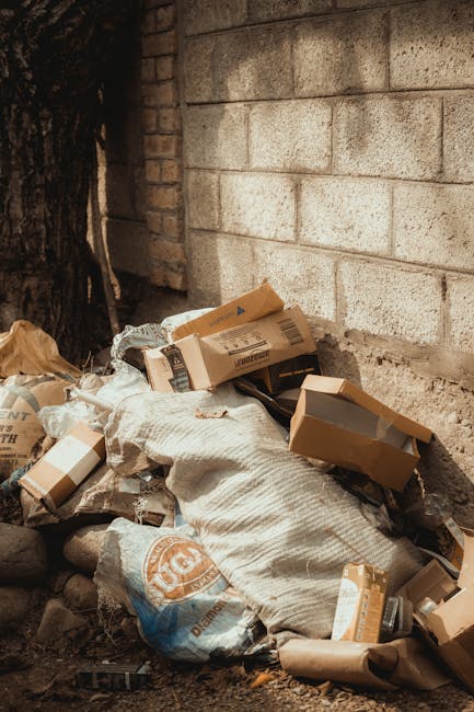 A pile of mixed household rubbish is shown against a brick wall and a concrete block wall. The debris includes flattened cardboard boxes, some with printed labels and barcodes, and brown paper bags. There is also a large, white, textured sack partially covering the other waste materials. The cardboard boxes vary in size and are in shades of light brown, with some open and others sealed or partially collapsed. The setting appears to be an outdoor area, possibly a driveway or yard, with the waste placed directly on uneven ground that has small stones and soil, and near the base of a tree trunk on the left side of the image. The scene suggests a collection of non-commercial domestic waste awaiting removal, aligning with private rubbish removal services such as those offered by Waste Removal Colliers Wood. The lighting is natural, casting soft shadows, and highlights the textures of the cardboard, paper, and rough brick and concrete surfaces, emphasizing the informal and unmanaged state of the waste accumulation.