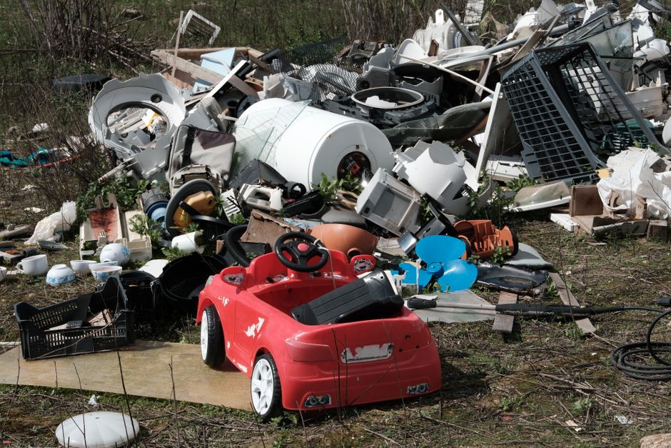 An outdoor scene showing several overflowing rubbish bins and scattered waste on a paved area adjacent to a parking lot. The central grey bin is open at the top, filled with mixed paper, cardboard, and plastic waste. To its right, there are black and red bins, with black bin bags and loose trash spilling onto the ground, including cardboard boxes, plastic packaging, and paper waste. The scene is set in front of a commercial building with a blue scaffolding structure on the upper level and storefronts with yellow signage below, indicating retail shops or services. On the left, a parked silver car is visible behind a metal railing, with a leafless tree nearby. The environment suggests a public space where waste has accumulated, highlighting the importance of proper rubbish collection and disposal services such as those offered by Waste Removal Colliers Wood, especially for efficient on-site clearance and alternative waste handling in urban areas.