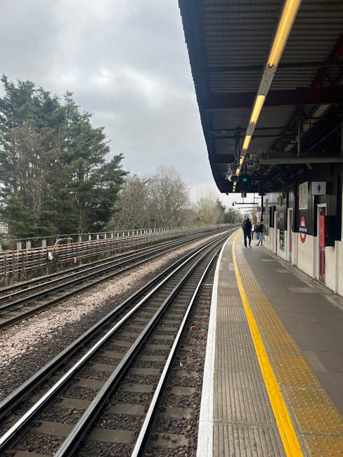 The image depicts a railway station platform adjacent to a set of train tracks, with a curved section of the tracks extending into the distance. The platform surface is made of concrete with a tactile yellow strip along the edge for safety, and features a roof overhead supported by metal beams, with lighting fixtures attached to the underside of the roof. Two people are walking along the platform, positioned near the center of the image, with their backs turned toward the camera. Behind them, the platform continues towards the horizon, bordered by a fence, with dense trees visible in the background beyond the tracks. The weather appears overcast, with gray clouds filling the sky, creating diffuse lighting conditions. To the right, adjacent to the platform, there are small structures including a ticket machine and an information board, indicating this is a public transportation setting. The scene is clean and organized, aligning with typical images associated with railway infrastructure, while also subtly connecting to the context of waste management and disposal in public areas such as train stations, as handled by services like Waste Removal Colliers Wood.