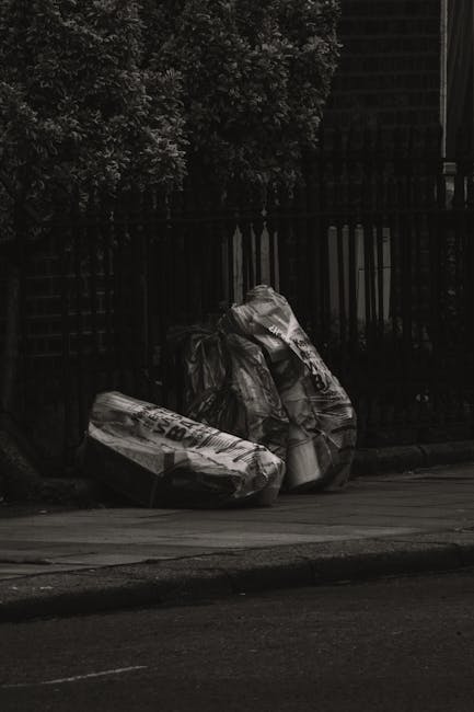 Two large bags of waste sit on a paved sidewalk in front of a black metal fence with vertical bars and a louvered gate, surrounded by dark foliage from trees nearby. The bags appear to be made of thick plastic material, with one partially open, revealing light-colored contents inside. The scene is illuminated by natural lighting, with shadows cast on the pavement and fence, indicating an outdoor setting possibly in the early morning or late afternoon. The bags contain household rubbish and are positioned close to the curb, suggesting they are awaiting collection or disposal. This image exemplifies private waste handling, often managed by local waste removal services such as Waste Removal Colliers Wood, who provide alternative rubbish collection options outside of standard council services.