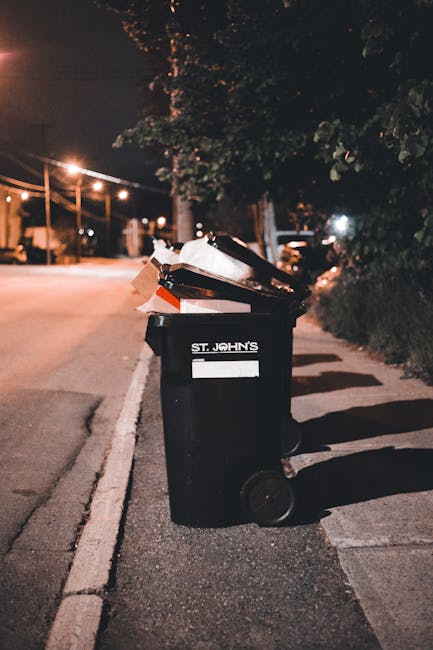 A black plastic wheeled rubbish bin positioned on the curbside of a paved street at night, with several open and partially filled cardboard boxes and loose waste piled on top. The bin is located in a residential area, with a row of trees and dense shrubbery on the right side, and a street illuminated by warm streetlights in the background. The bin has a white label with the name 'St. John's' and is situated close to a paved sidewalk with a concrete curb. The scene shows a quiet urban setting, possibly indicating private waste disposal or alternative waste handling outside of regular collection times, reflecting services offered by Waste Removal Colliers Wood in the context of rubbish and waste management. The overall atmosphere is dimly lit, with shadows cast on the ground and a subtle glow from the streetlights, emphasizing the bin’s role in waste collection at night.